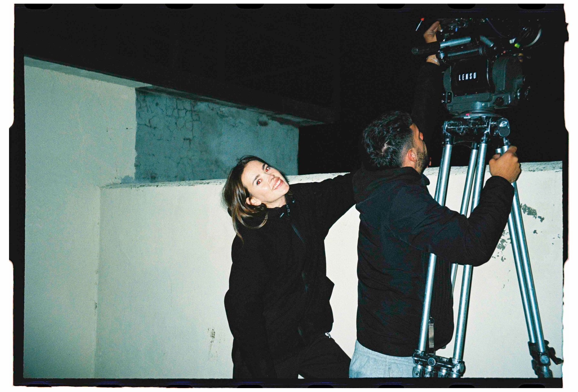Directora sonriendo junto al operador de cámara ajustando el trípode Lenso en el set nocturno, 35mm